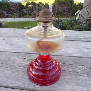 May include: A vintage oil lamp with a clear glass reservoir and a red glass base. The lamp has a metal top and a wick visible inside. The lamp is sitting on a weathered wooden surface.