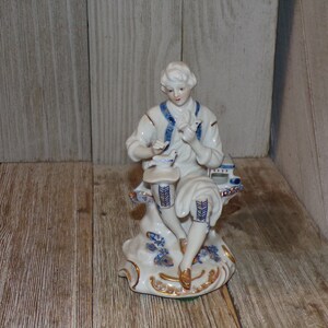 Pu&ograve; includere: Una statuetta in porcellana di una persona seduta su una panchina, che tiene un uccello in mano. La statuetta &egrave; dipinta in bianco, blu e oro.