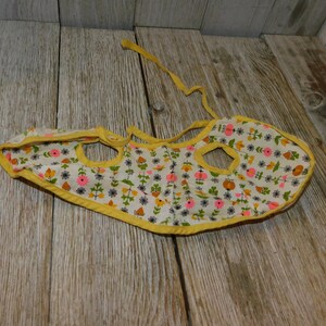 May include: A yellow and white floral print baby bib with a yellow trim and two openings for arms. The bib has a tie at the back.