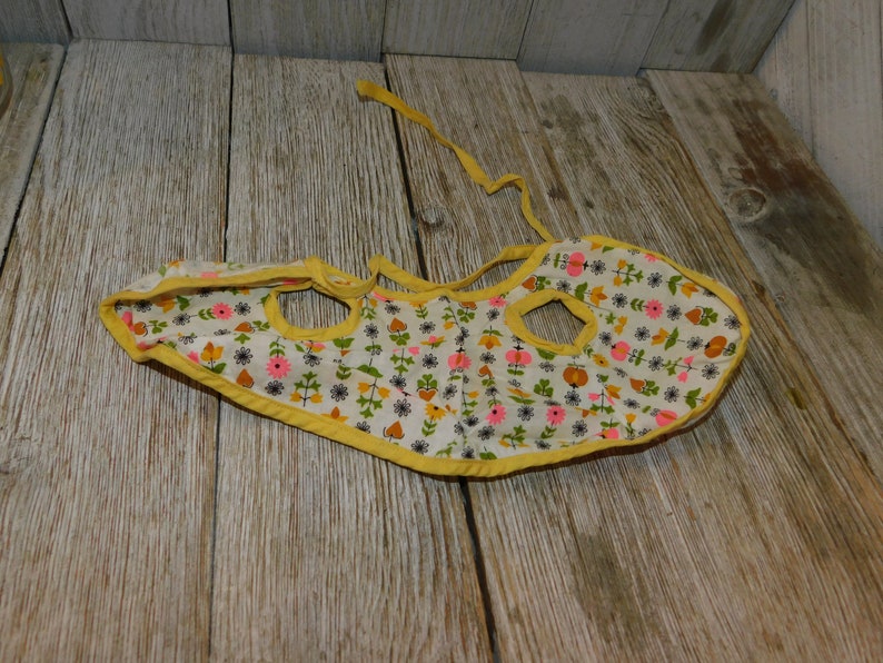 May include: A yellow and white floral print baby bib with a yellow trim and two openings for arms. The bib has a tie at the back.