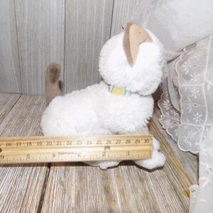 May include: A white plush cat toy with tan ears and tail, wearing a colorful collar. A wooden ruler is placed next to the toy. The cat toy measures approximately 10 inches in length.