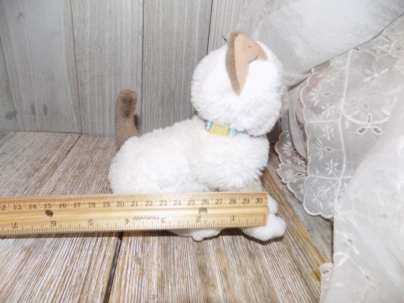 May include: A white plush cat toy with tan ears and tail, wearing a colorful collar. A wooden ruler is placed next to the toy. The cat toy measures approximately 10 inches in length.