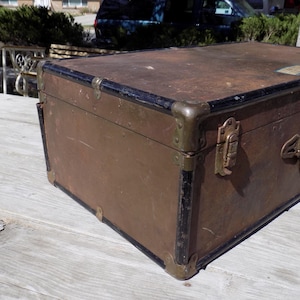 May include: A vintage brown trunk with black trim and brass hardware. The trunk has a rectangular shape with metal corner protectors and a handle. The trunk is likely used for storage or travel.