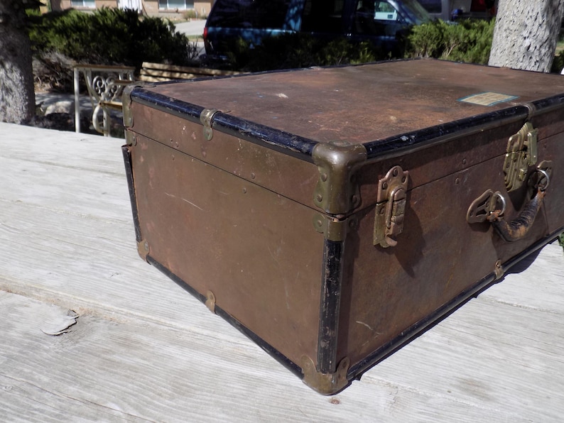 May include: A vintage brown trunk with black trim and brass hardware. The trunk has a rectangular shape with metal corner protectors and a handle. The trunk is likely used for storage or travel.