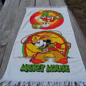 Peut inclure: Serviette rectangulaire blanche avec une illustration color&eacute;e de Mickey Mouse en tenue de cow-boy, tenant un lasso. La serviette a un bord &agrave; franges et les mots "MICKEY MOUSE" en vert.
