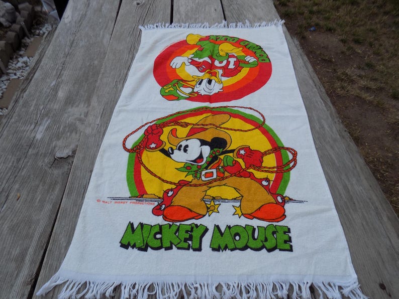Peut inclure: Serviette rectangulaire blanche avec une illustration color&eacute;e de Mickey Mouse en tenue de cow-boy, tenant un lasso. La serviette a un bord &agrave; franges et les mots "MICKEY MOUSE" en vert.