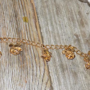 May include: Gold-coloured charm bracelet with several charms. The charms depict Mickey Mouse and other Disney characters. The bracelet is made of linked chain and is shown on a wooden surface.