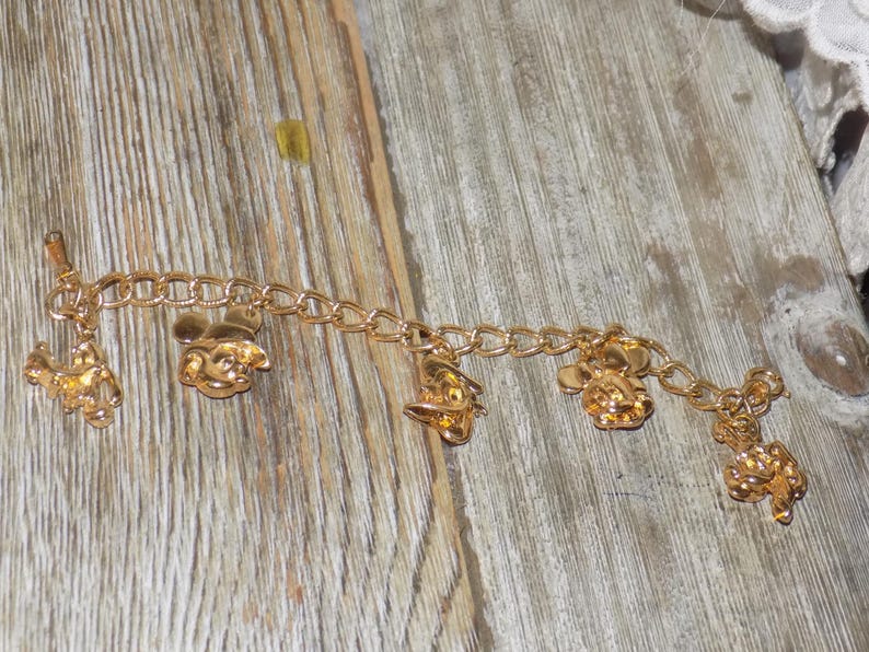 May include: Gold-coloured charm bracelet with several charms. The charms depict Mickey Mouse and other Disney characters. The bracelet is made of linked chain and is shown on a wooden surface.