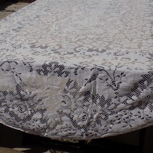 May include: A white lace tablecloth with a floral pattern covers a table. The lace has a delicate, openwork design, with detailed floral and geometric shapes. The tablecloth is draped over the table, showing its texture and intricate details.