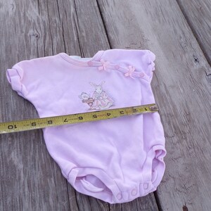 May include: A pink baby onesie with a bunny and a cat illustration. The onesie has pink bows and buttons on the shoulder. A measuring tape is placed across the onesie, showing a measurement of 10 inches.