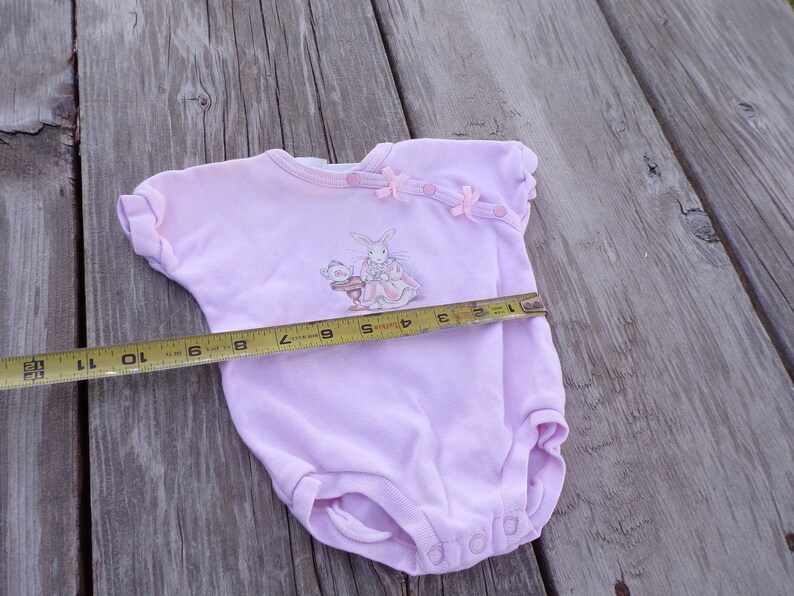 May include: A pink baby onesie with a bunny and a cat illustration. The onesie has pink bows and buttons on the shoulder. A measuring tape is placed across the onesie, showing a measurement of 10 inches.