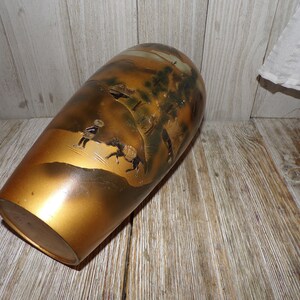 May include: A gold ceramic vase with a Japanese-inspired design featuring a landscape scene with figures and trees. The vase has a slightly rounded shape and a smooth, glossy finish.