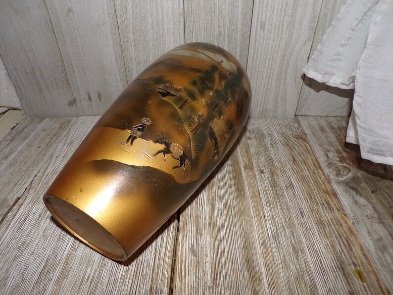 May include: A gold ceramic vase with a Japanese-inspired design featuring a landscape scene with figures and trees. The vase has a slightly rounded shape and a smooth, glossy finish.
