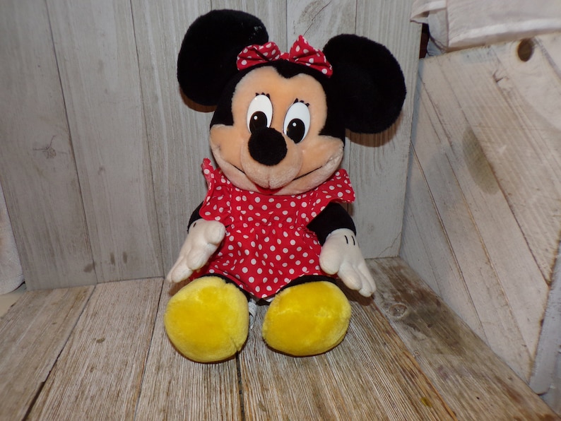 K&ouml;nnte beinhalten: Ein Pl&uuml;schtier von Minnie Mouse, die ein rotes Kleid mit wei&szlig;en Punkten und eine rote Schleife tr&auml;gt. Sie hat schwarze Ohren und eine schwarze Nase.