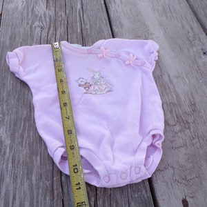 May include: A pink baby onesie with a bunny rabbit graphic and pink bows. The onesie is being measured with a tape measure.