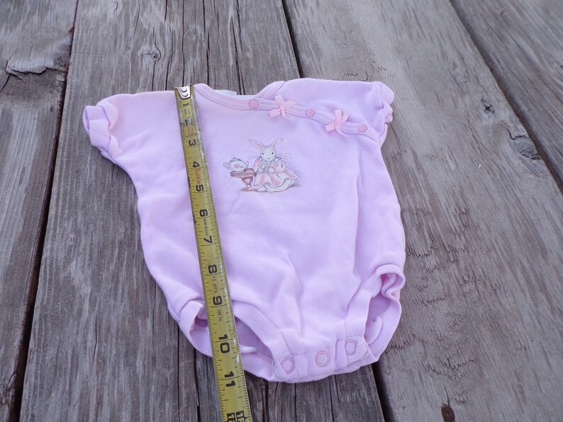 May include: A pink baby onesie with a bunny rabbit graphic and pink bows. The onesie is being measured with a tape measure.