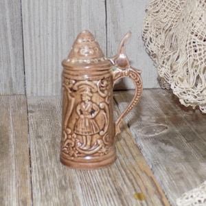 May include: A brown ceramic beer stein with a lid and a handle. The stein is decorated with a raised design of a woman in a dress and flowers.