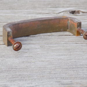 May include: A weathered brass drawer pull with a rectangular arch shape and a dull, tarnished finish. The hardware includes two integrated metal screws with visible oxidation and rust, extending outward from the ends of the handle base.