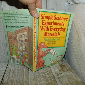 May include: A book titled "Simple Science Experiments With Everyday Materials" by Muriel Mandell. The cover features a green background with a yellow banner and illustrations of a cartoon dinosaur and a frog. The book is held by a hand.