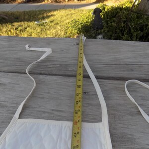 Op de afbeelding: Witte katoenen schort met twee banden en een meetlint dat een lengte van 45,7 cm aangeeft.