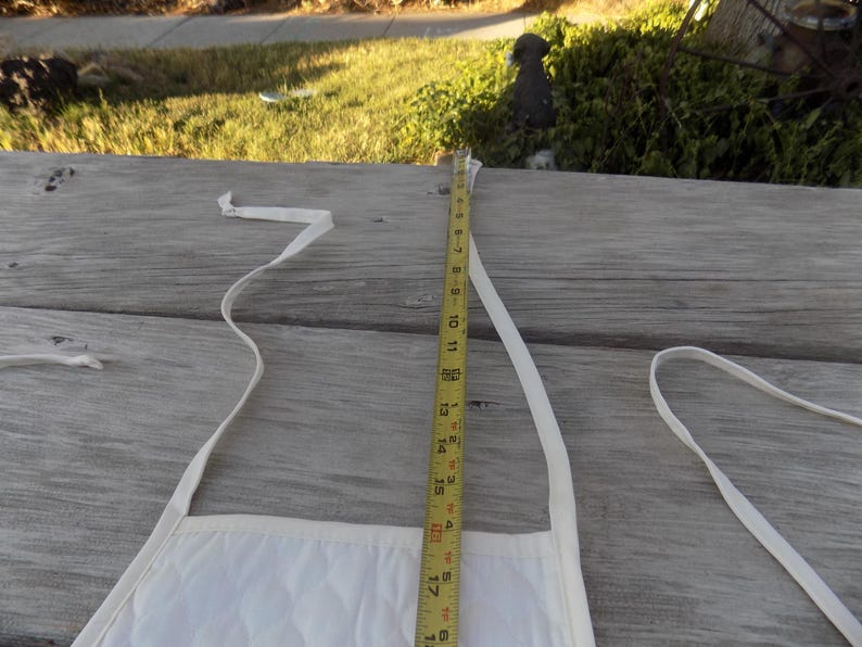 Op de afbeelding: Witte katoenen schort met twee banden en een meetlint dat een lengte van 45,7 cm aangeeft.