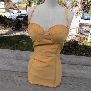 May include: Mustard yellow one-piece swimsuit with a sweetheart neckline and thin shoulder straps. The swimsuit is displayed on a mannequin. The garment has a fitted silhouette and is made of a smooth fabric.
