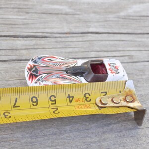 Puede incluir: Un coche de carreras de juguete blanco con gr&aacute;ficos de llamas rojas, negras y plateadas y una cabina marr&oacute;n oscuro. Una cinta m&eacute;trica amarilla est&aacute; colocada al lado, mostrando que mide aproximadamente 7 cm desde el parachoques hasta atr&aacute;s.