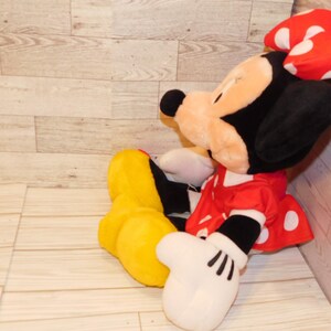 Op de afbeelding: Een Minnie Mouse knuffel, met een zwarte kop en oren, een rode jurk met witte stippen en een rode strik met witte stippen. De pop heeft witte handschoenen en gele schoenen.