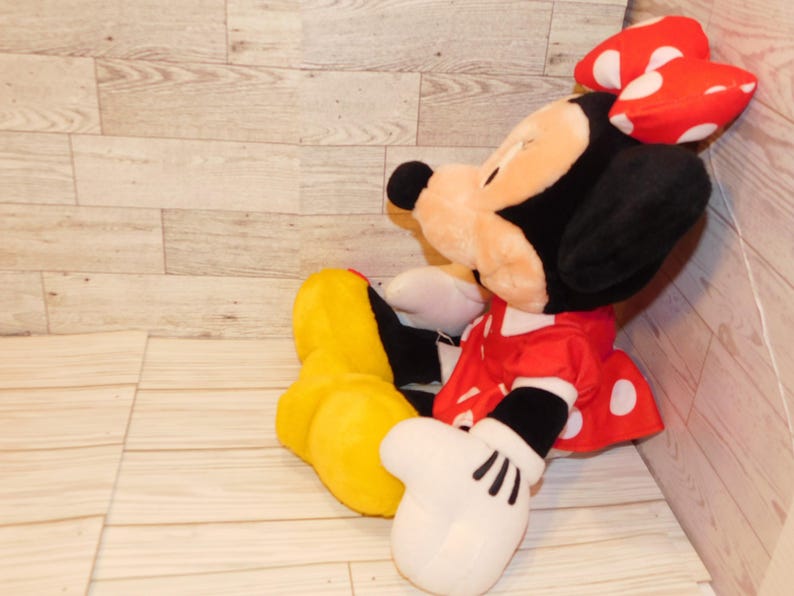 Op de afbeelding: Een Minnie Mouse knuffel, met een zwarte kop en oren, een rode jurk met witte stippen en een rode strik met witte stippen. De pop heeft witte handschoenen en gele schoenen.