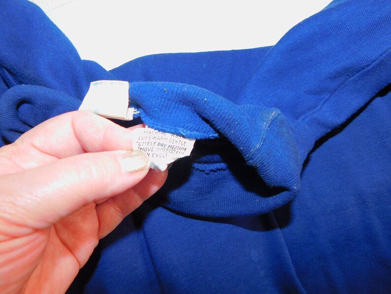 May include: Close-up of a blue fabric care label with the text "Machine Wash Lukewarm Gentle Tumble Dry Medium Move Immediately From Dryer To Prevent Wrinkles"