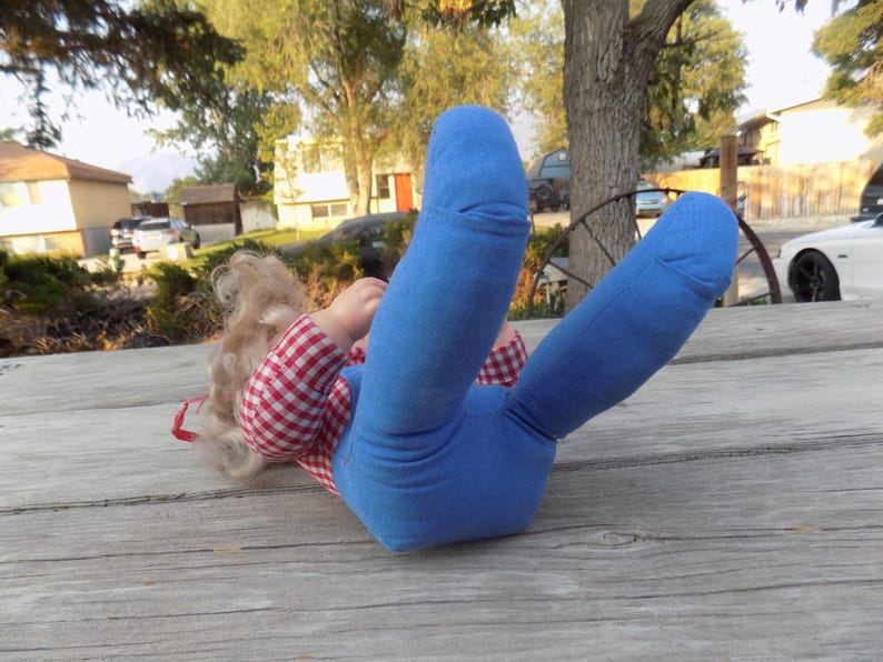 Puede incluir: Una mu&ntilde;eca con una camisa a cuadros rojos y blancos y pantalones vaqueros azules. La mu&ntilde;eca tiene el pelo rubio y est&aacute; tumbada sobre una superficie de madera.