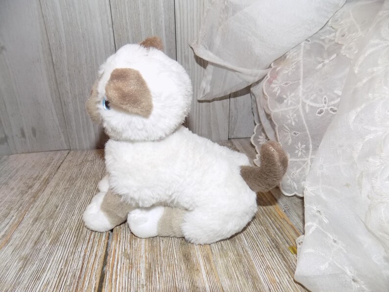 May include: A plush white and brown cat toy with blue eyes. The stuffed animal is sitting on a wooden surface. The cat has brown ears, paws, and tail. The toy is soft and fluffy.