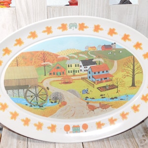 May include: Oval serving platter with a rural autumn scene. The white rim is decorated with orange fall leaves. The center features a village with houses, a barn, a water wheel, and a stream. The platter is a decorative piece.