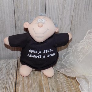 May include: A plush doll with a smiling face, gray hair, and a black shirt. The shirt has the text "ONCE A STUD... ALWAYS A STUD" in white. The doll has a light skin tone and is standing on a wooden surface.