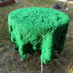May include: A round table covered with a bright green, hand-crocheted tablecloth. The tablecloth has a lacy, openwork design and hangs down the sides of the table. The table has a white metal frame and is set on a grassy surface.