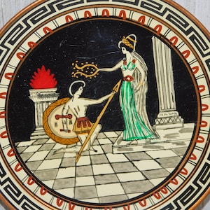Pu&ograve; includere: Un piatto rotondo in rame con sfondo nero e una scena dipinta di un uomo e una donna in un ambiente greco. Il piatto ha un bordo decorativo con un motivo a chiave greca in bianco e nero.