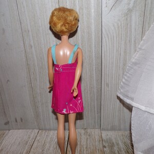 May include: A vintage Barbie doll wearing a pink dress with a floral print and blue straps. The doll has blonde hair styled in a short bob.