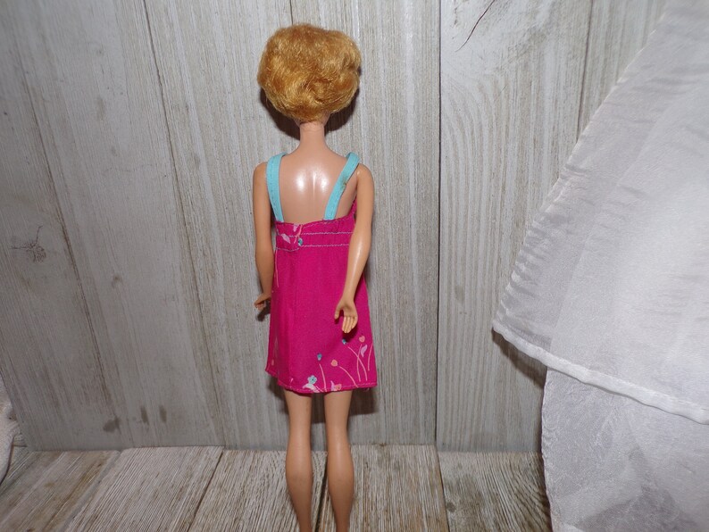 May include: A vintage Barbie doll wearing a pink dress with a floral print and blue straps. The doll has blonde hair styled in a short bob.