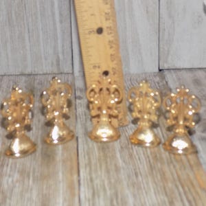 May include: Five gold-toned metal place card holders with a decorative design. Each holder has a round base and a decorative top.