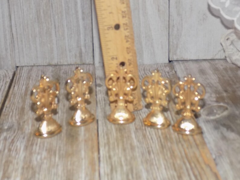 May include: Five gold-toned metal place card holders with a decorative design. Each holder has a round base and a decorative top.