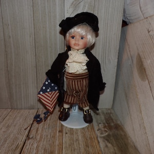 May include: A porcelain doll dressed in a black coat, white shirt, and striped pants, holding a small American flag. The doll is wearing a black hat and has blonde hair. The doll is standing on a white stand.