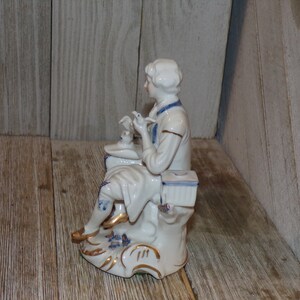 Pu&ograve; includere: Una statuetta in porcellana di una persona seduta su una base, che tiene un uccello in mano. La statuetta &egrave; dipinta in blu e bianco con accenti dorati.