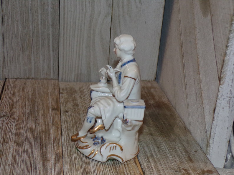Pu&ograve; includere: Una statuetta in porcellana di una persona seduta su una base, che tiene un uccello in mano. La statuetta &egrave; dipinta in blu e bianco con accenti dorati.
