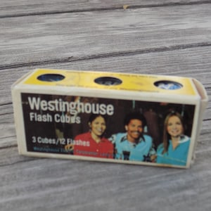 May include: A rectangular Westinghouse Flash Cubes box featuring a graphic of three people and the text Westinghouse Flash Cubes 3 Cubes/12 Flashes. The yellow top panel has three circular cutouts revealing the blue flash cubes inside the packaging.