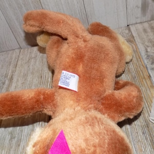 May include: A brown plush toy rabbit with long ears and a pink diamond-shaped accent on its belly. The rabbit has a tag on its back with text. The toy is lying on a wooden surface.