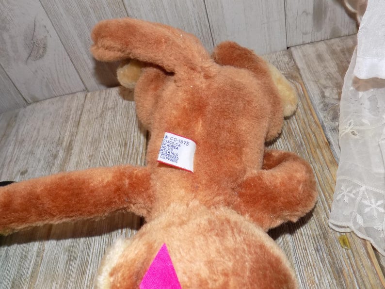 May include: A brown plush toy rabbit with long ears and a pink diamond-shaped accent on its belly. The rabbit has a tag on its back with text. The toy is lying on a wooden surface.
