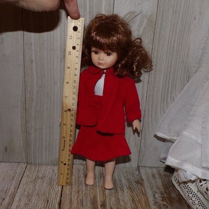 K&ouml;nnte beinhalten: Eine Porzellanpuppe mit braunen Haaren, die ein rotes Kleid und eine rote Jacke tr&auml;gt. Die Puppe ist etwa 30 cm gro&szlig;.