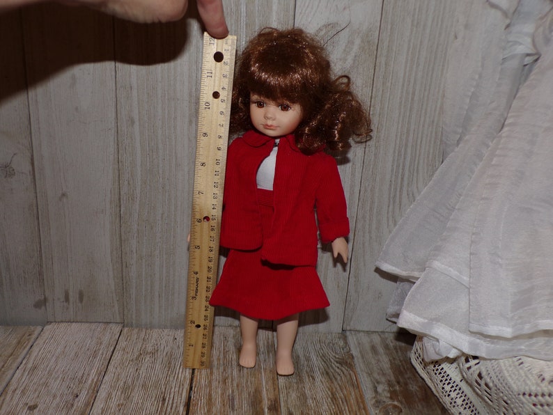 K&ouml;nnte beinhalten: Eine Porzellanpuppe mit braunen Haaren, die ein rotes Kleid und eine rote Jacke tr&auml;gt. Die Puppe ist etwa 30 cm gro&szlig;.