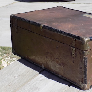 May include: An antique trunk with a weathered, brown exterior and black trim. The trunk features metal corner protectors and hinges, suggesting a vintage travel or storage item. The aged appearance indicates it has been well-used over time.