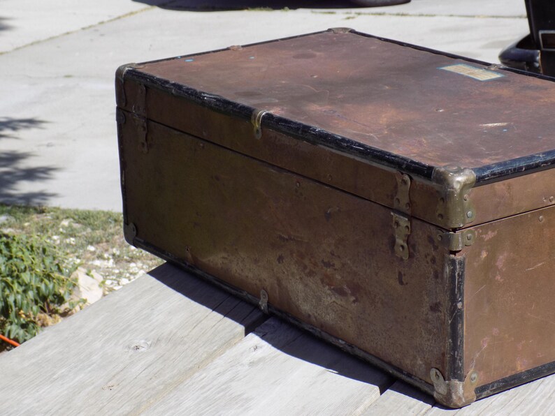 May include: An antique trunk with a weathered, brown exterior and black trim. The trunk features metal corner protectors and hinges, suggesting a vintage travel or storage item. The aged appearance indicates it has been well-used over time.
